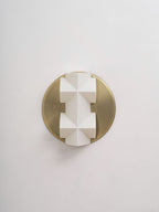 Close, straight-on view of an unlit white faceted Clérachic wall sconce on a round brushed gold backplate, on a pale wall.