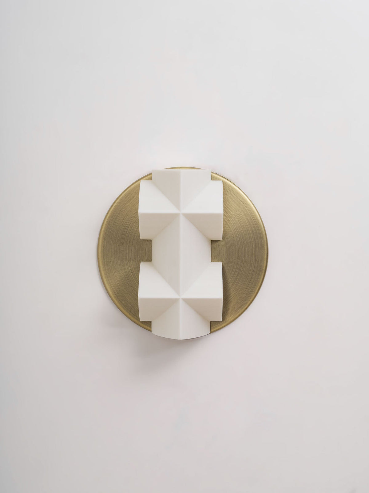 Close, straight-on view of an unlit white faceted Clérachic wall sconce on a round brushed gold backplate, on a pale wall.