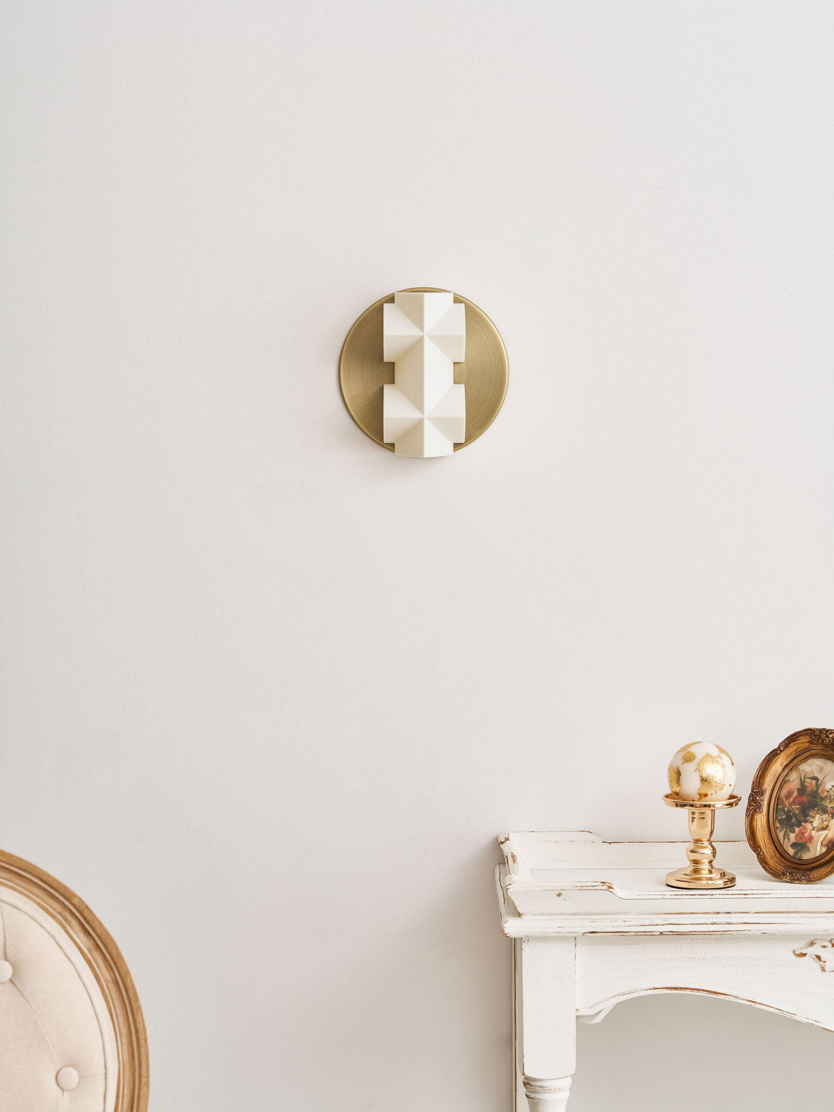 A small unlit Clérachic wall sconce on a gold disc, high on a pale wall above a white console with a gilt globe.