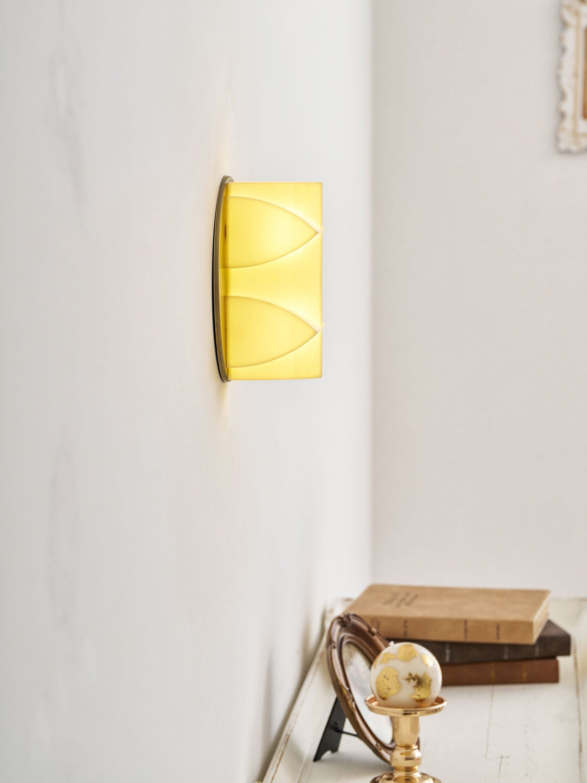 Side-on view of a Clérachic wall sconce lit a soft yellow, with books and a gilt ornament on a shelf below.