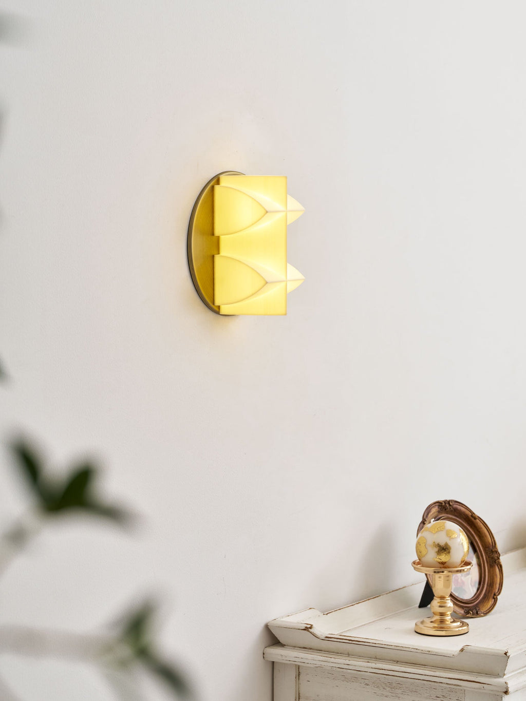 Angled view of a lit Clérachic sconce on a pale wall, its curved points glowing, above a white table with a gilt ornament.