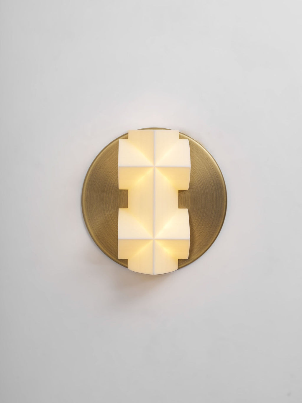A lit Clérachic wall sconce seen straight on, its faceted white shade glowing warm on a round gold plate, on a pale grey wall.