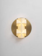 A lit Clérachic wall sconce seen straight on, its faceted white shade glowing warm on a round gold plate, on a pale grey wall.