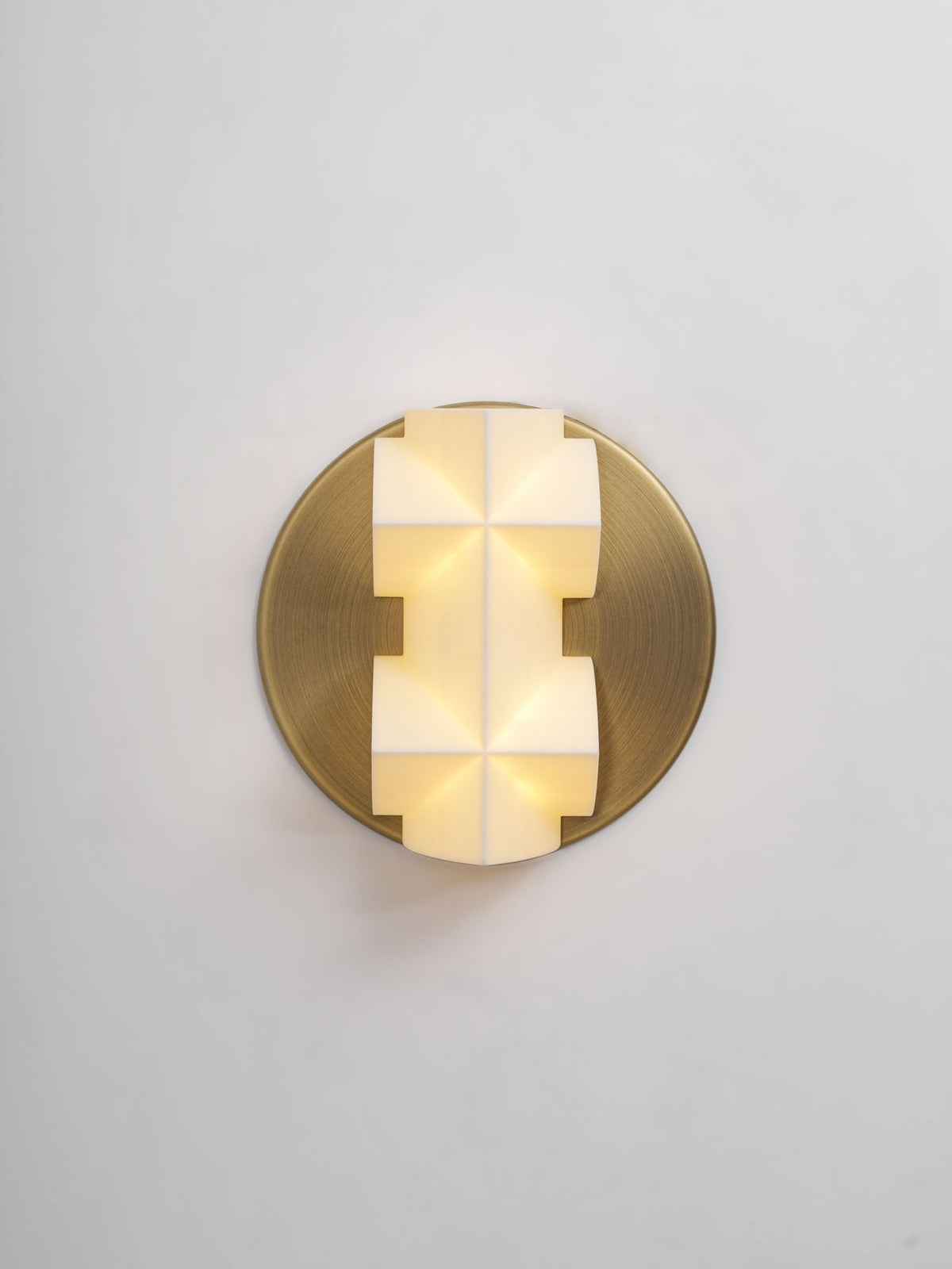 A lit Clérachic wall sconce seen straight on, its faceted white shade glowing warm on a round gold plate, on a pale grey wall.