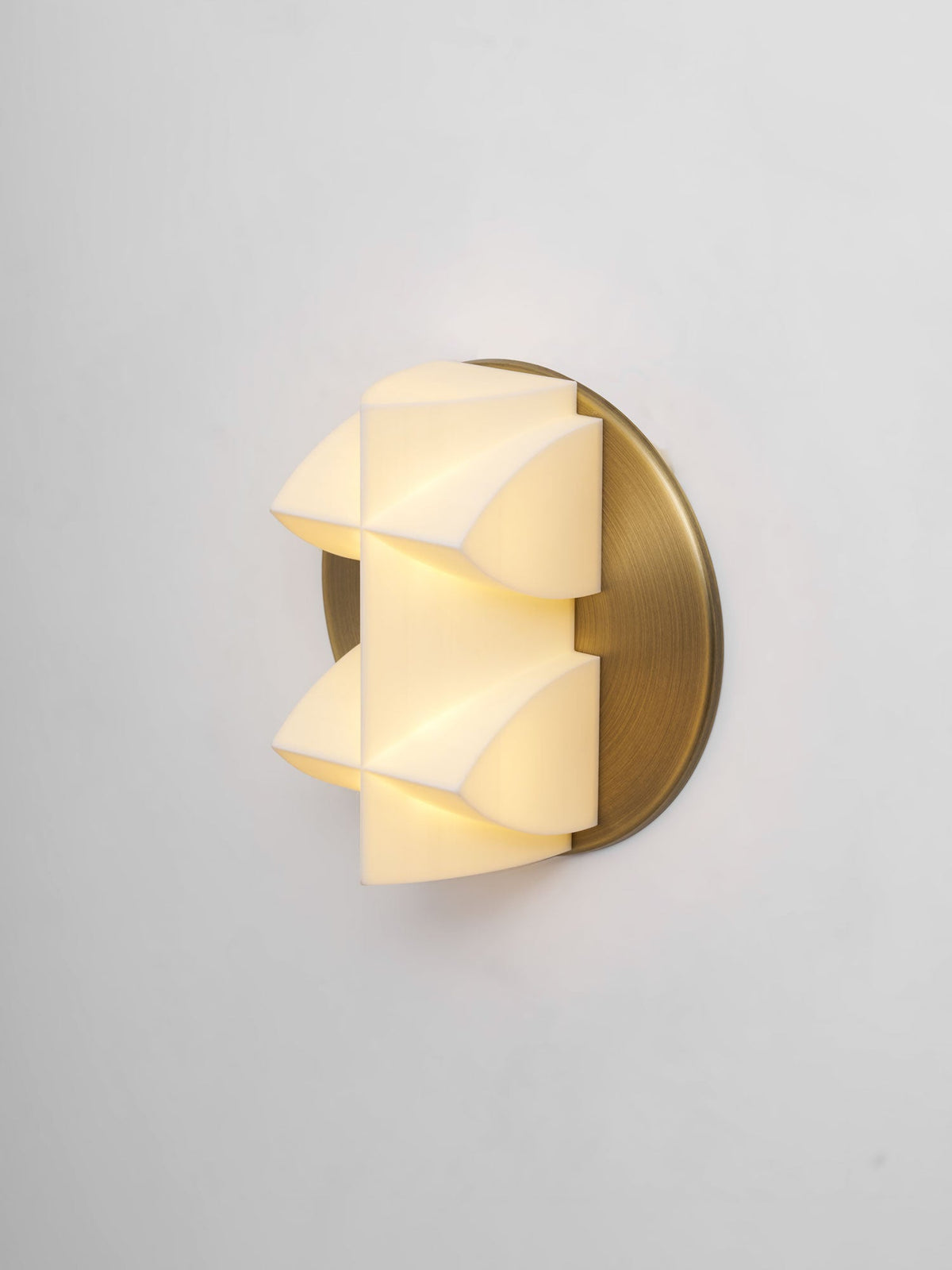Lit Clérachic sconce seen at an angle, warm light spilling through the white facets beside a gold backplate.
