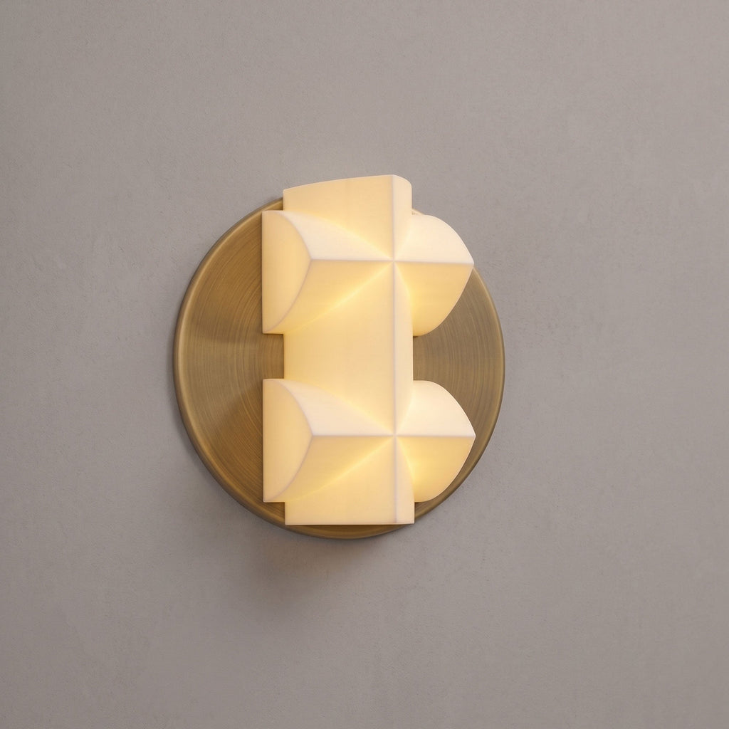 A lit Clérachic wall sconce in close view, its faceted white shade on a gold disc, against a taupe wall.
