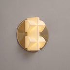 A lit Clérachic wall sconce in close view, its faceted white shade on a gold disc, against a taupe wall.