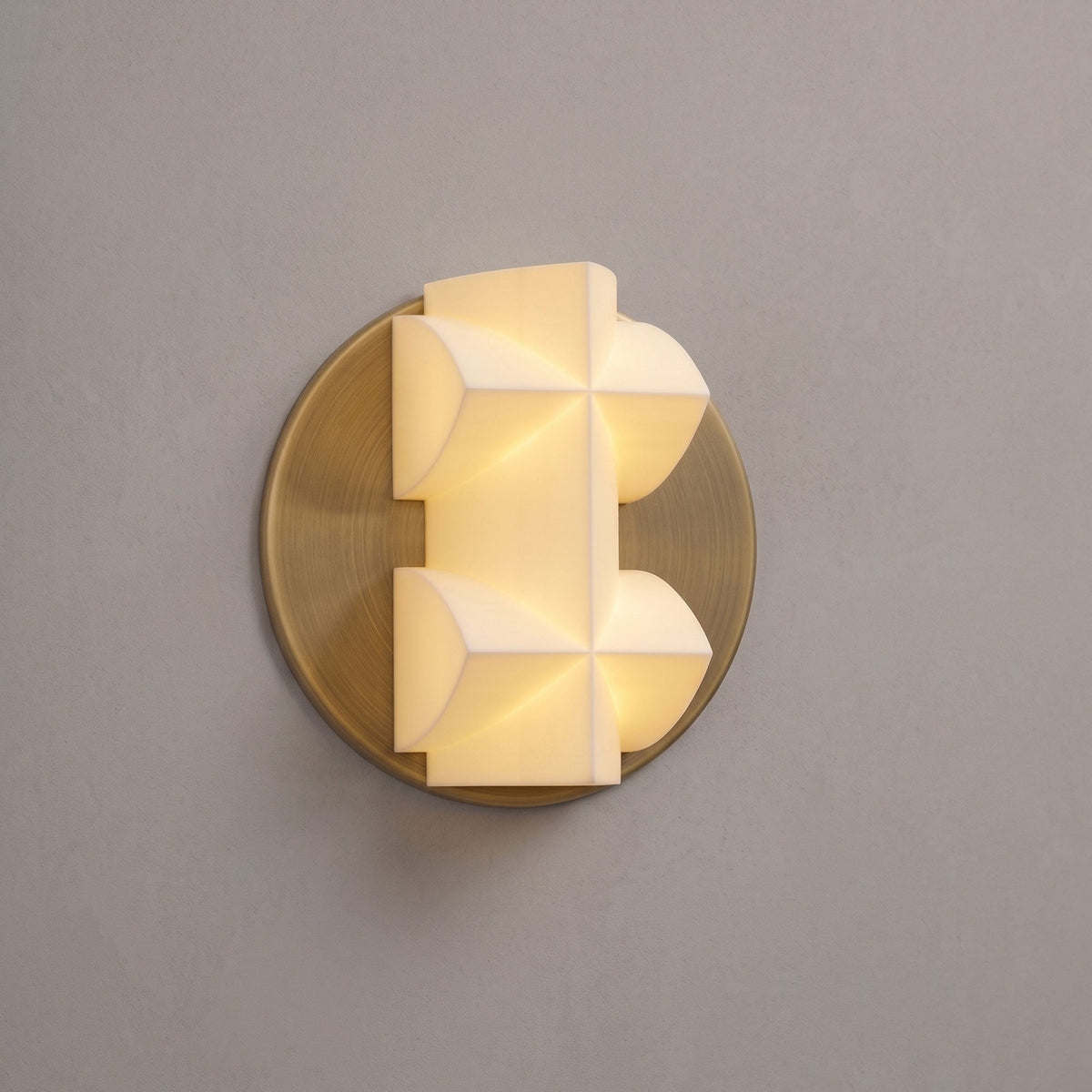 A lit Clérachic wall sconce in close view, its faceted white shade on a gold disc, against a taupe wall.