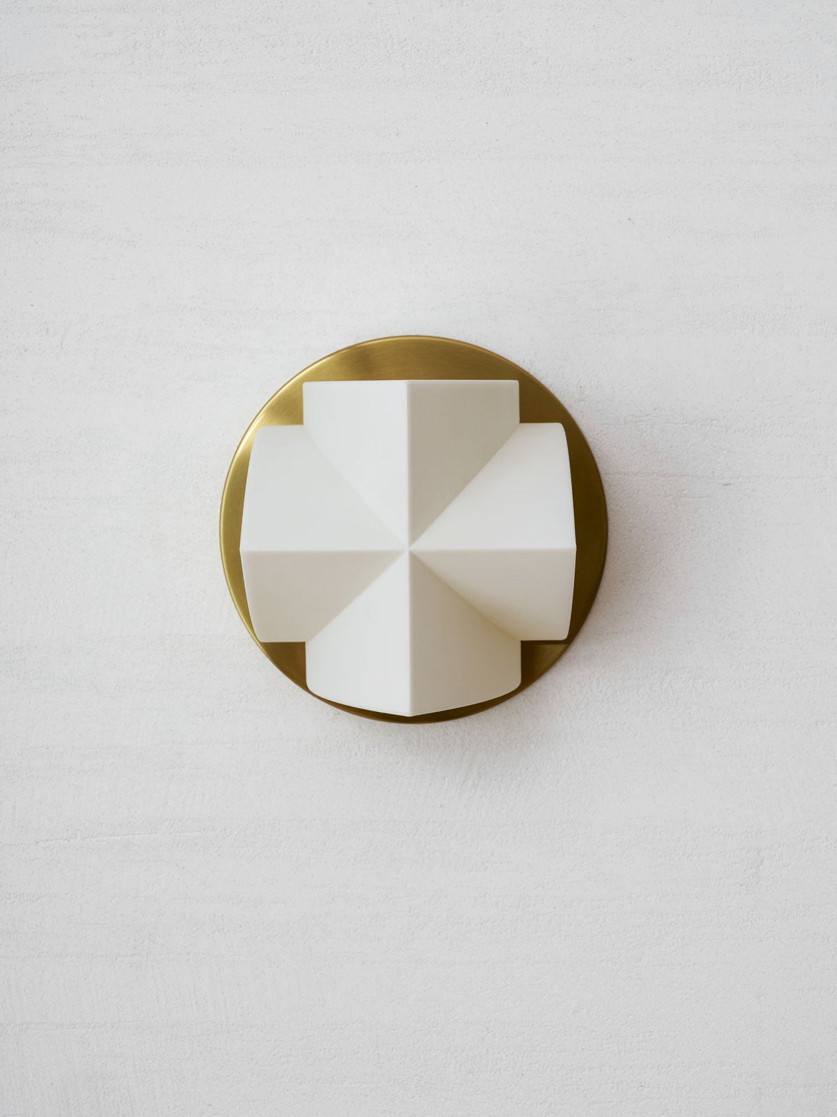 A single white crossed-facet shade, unlit, on a round gold backplate of this Clérachic sconce, on a textured white wall.