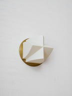 Unlit Clérachic wall sconce at an angle, its white facets forming a pointed shape in front of a brushed gold disc.