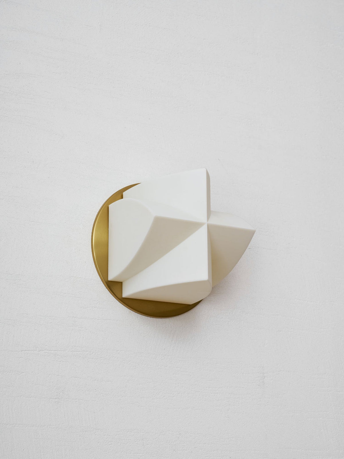 Unlit Clérachic wall sconce at an angle, its white facets forming a pointed shape in front of a brushed gold disc.