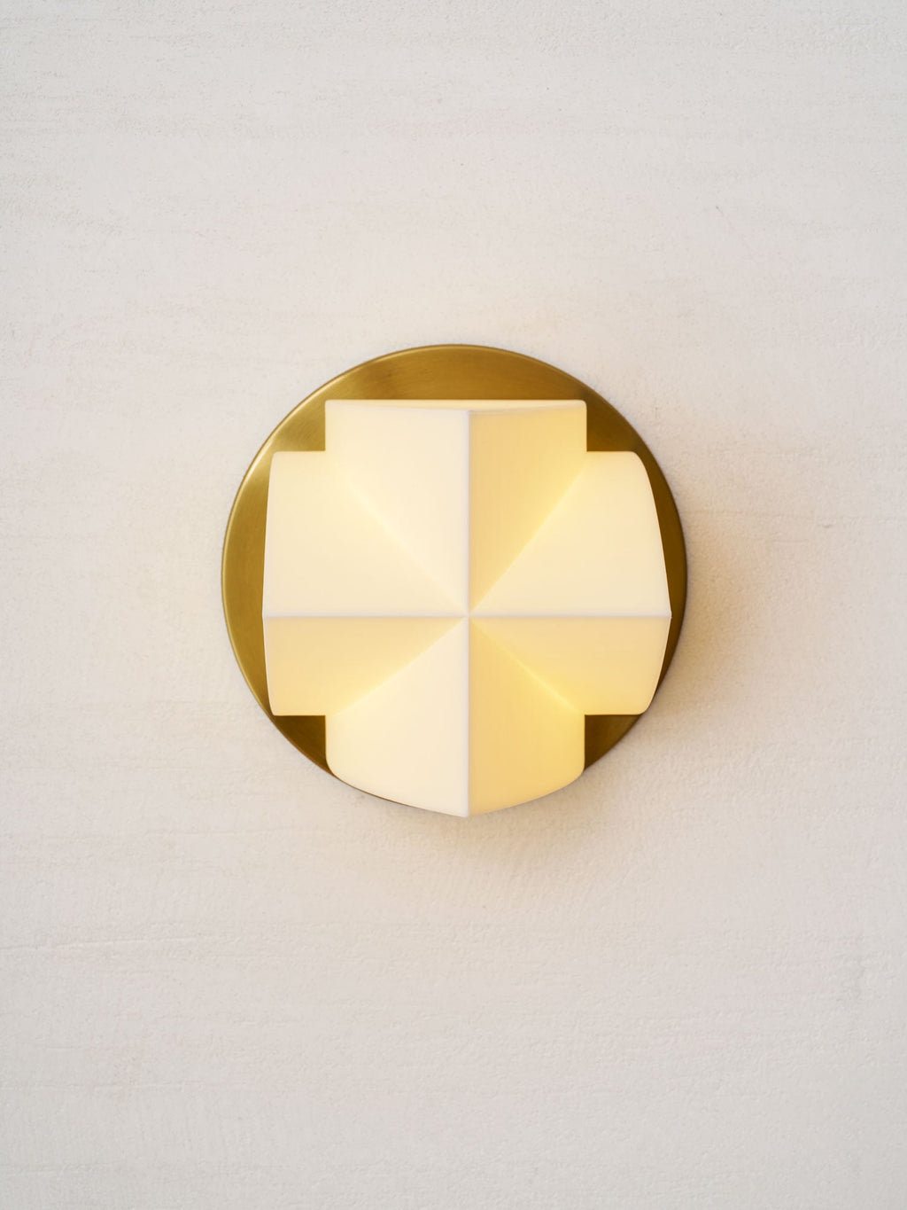 Front view of a lit Clérachic wall sconce, a cross of glowing white facets on a round gold plate, on a textured wall.