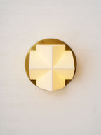 Front view of a lit Clérachic wall sconce, a cross of glowing white facets on a round gold plate, on a textured wall.
