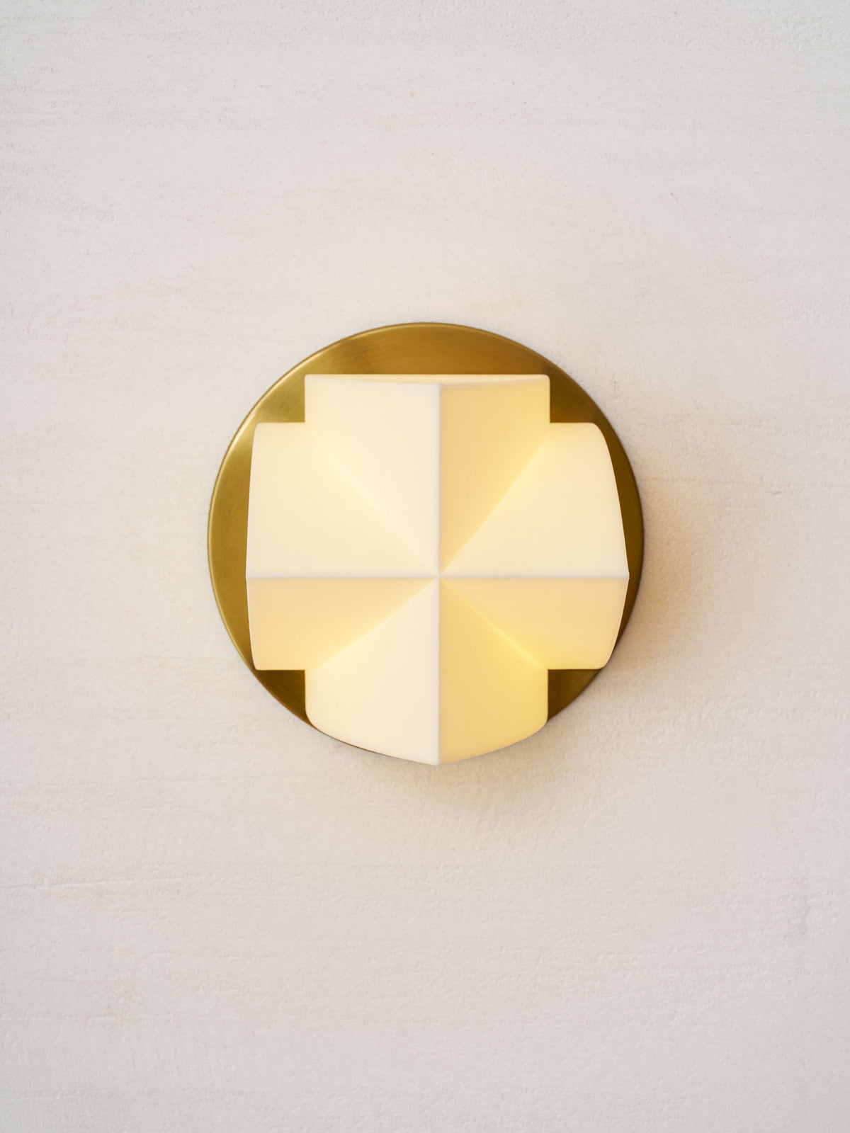 Front view of a lit Clérachic wall sconce, a cross of glowing white facets on a round gold plate, on a textured wall.