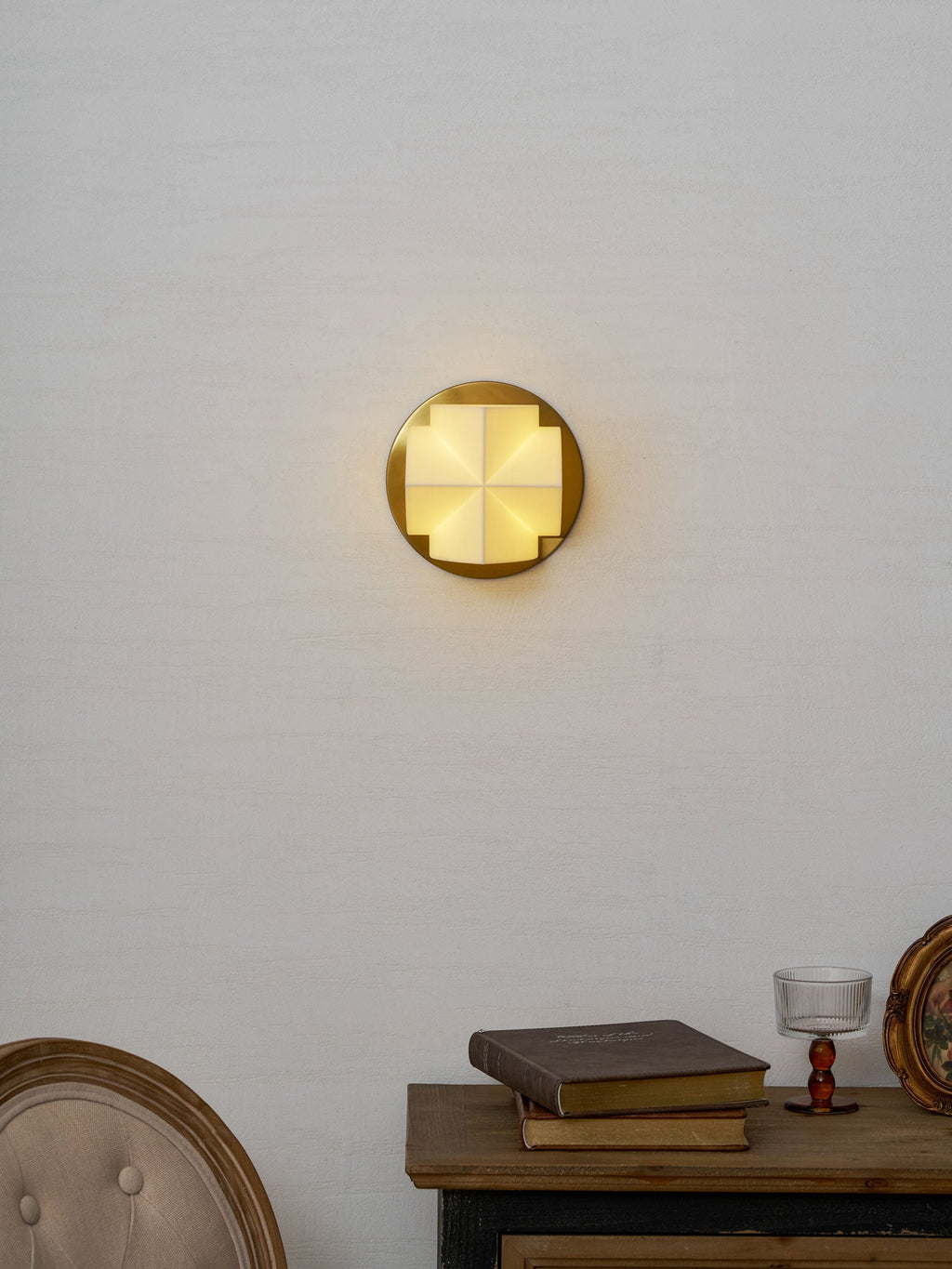 A Clérachic wall sconce lit warm and centred on a pale wall, above a console with books and a glass on a stem.