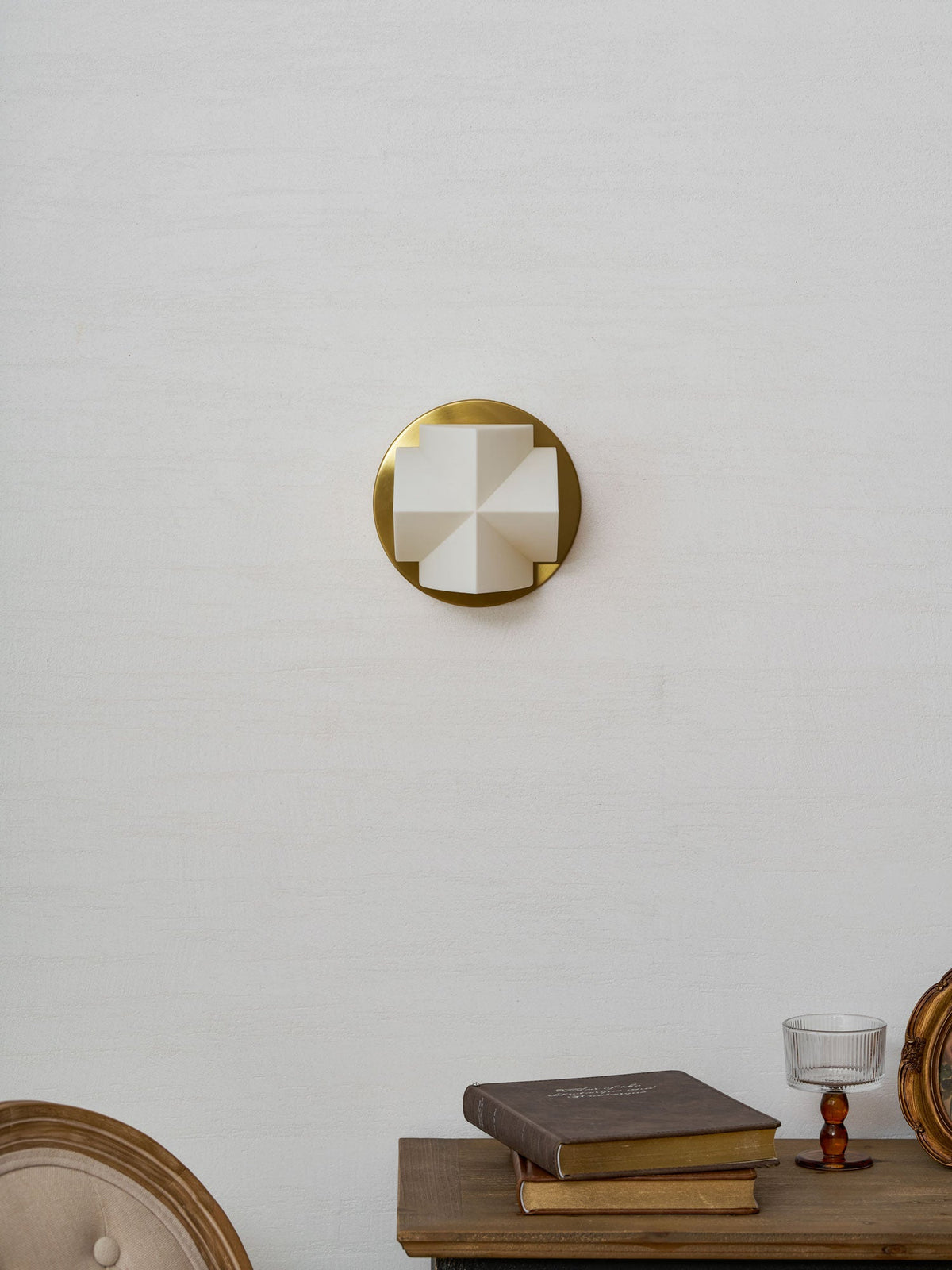 Unlit, a small Clérachic wall sconce shows its white crossed facets on a gold disc above a console with books.