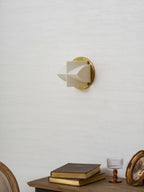 Side view of an unlit Clérachic wall sconce on a white wall, above a console with books, a glass and a round gilt frame.