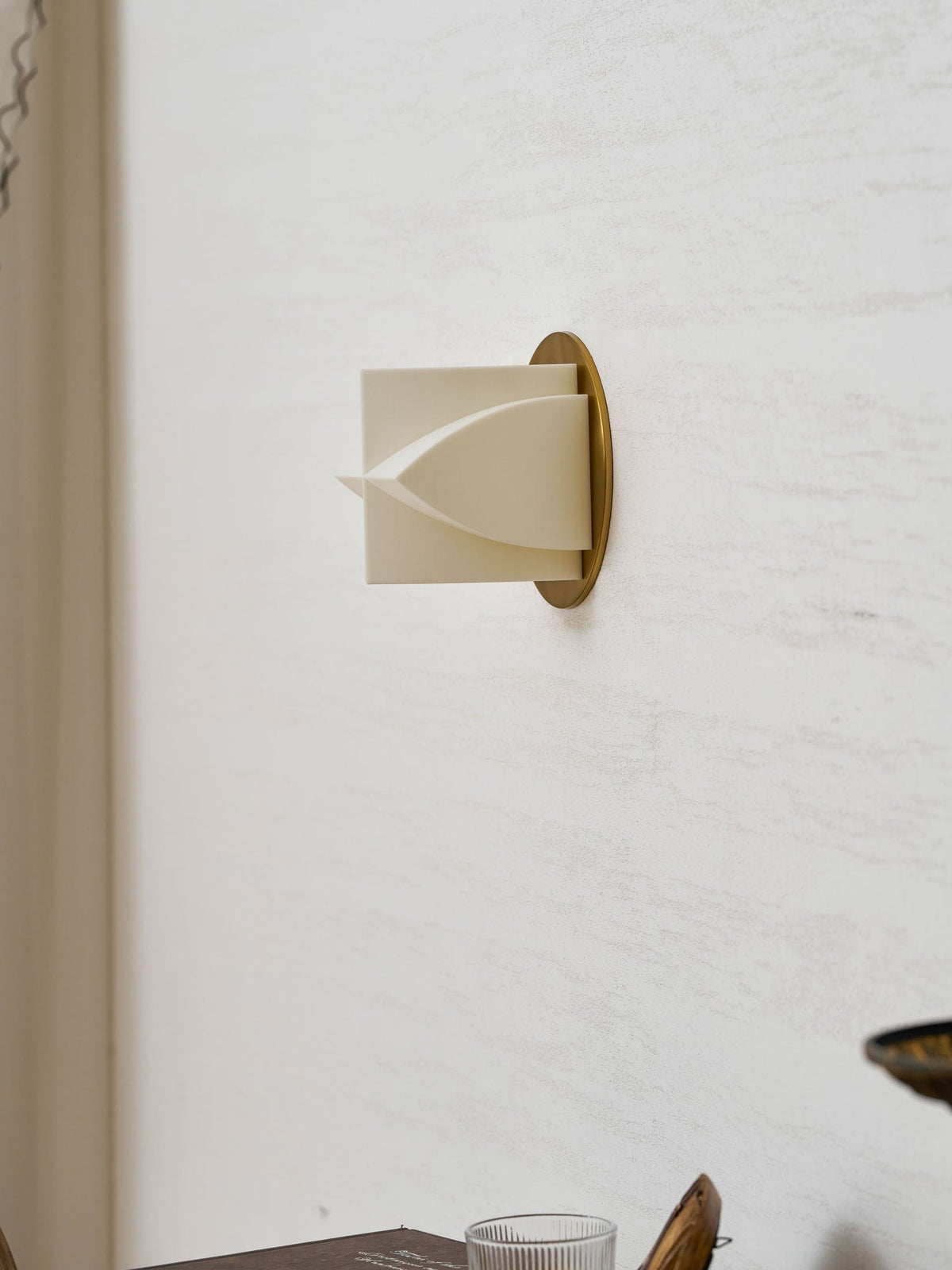 A Clérachic wall sconce in profile beside a door frame, its white facets pointing left from a gold disc.