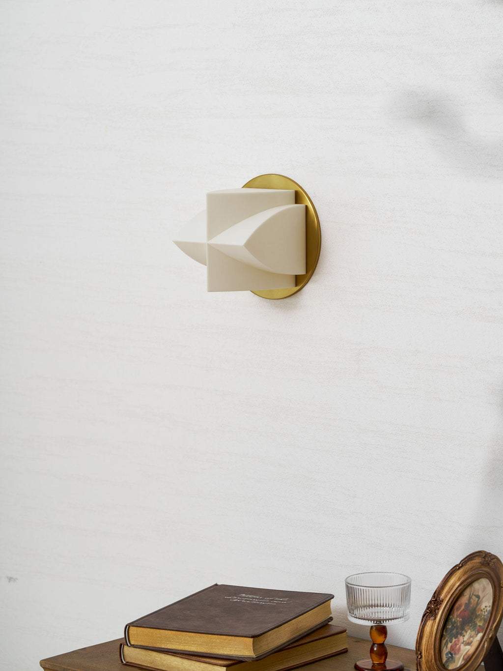 An unlit Clérachic sconce in profile on a white wall, with a stack of books and a round gilt frame below.