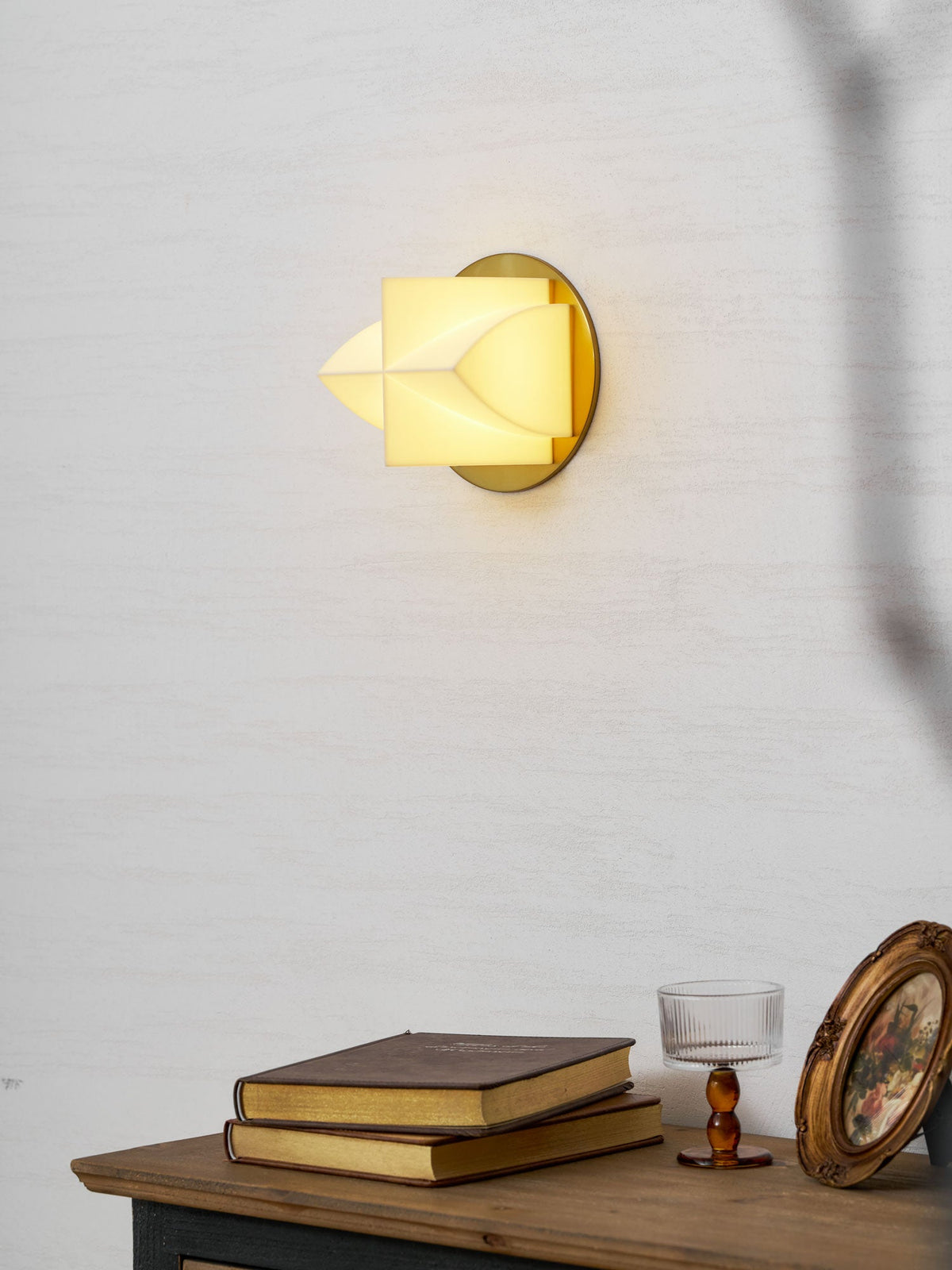 A Clérachic sconce lit yellow in profile, with a stack of books, a glass and a gilt frame on the console below.