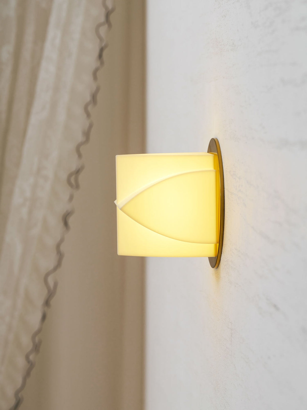 Close side view of a Clérachic wall sconce lit pale yellow, beside a lace curtain on a white wall.