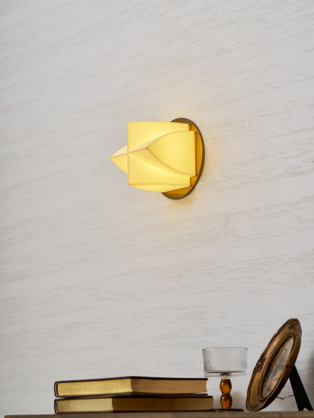 Lit Clérachic sconce in profile on a textured white wall, with books and a gilt frame on a console beneath it.