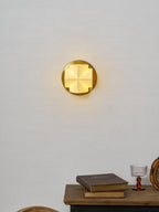 A small lit Clérachic sconce seen from the front, hung on a pale wall above a console with books and a glass.