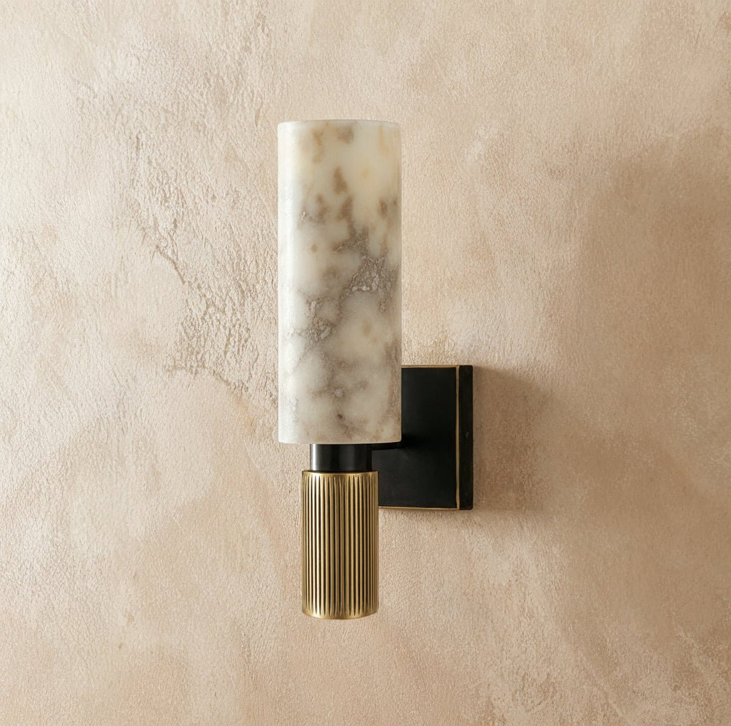 Clérachic gold wall sconce with a cylindrical alabaster stone shade and fluted brass base, mounted on a plaster wall.