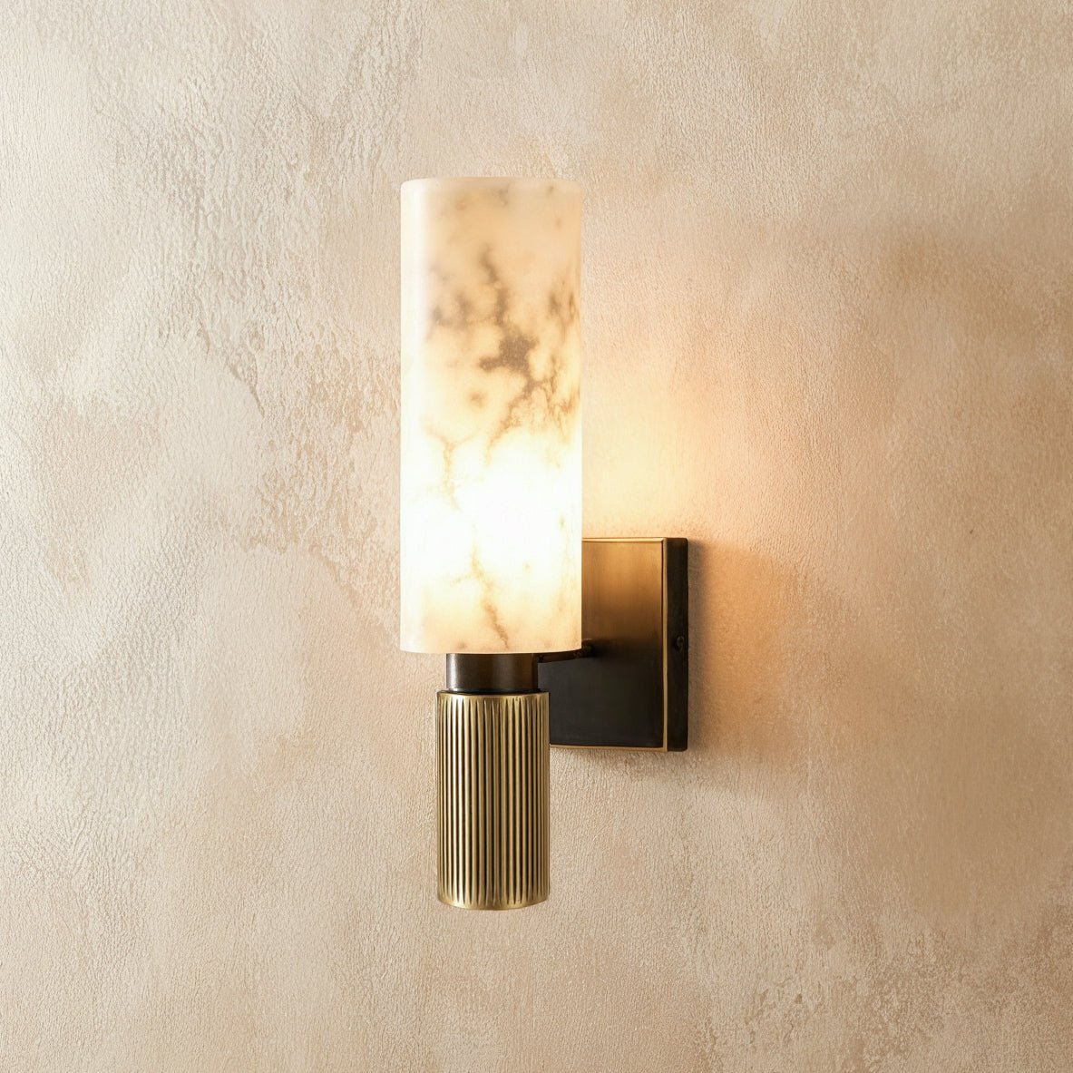 Clérachic alabaster wall sconce lit warmly, its veined stone cylinder glowing above a black-and-brass fitting.