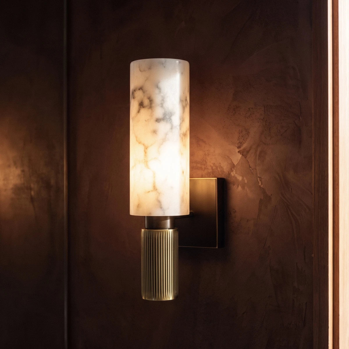 Clérachic gold alabaster wall sconce glowing against a deep rust-toned plaster wall near a timber door frame.