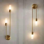 Two Clérachic gold stick sconces staggered on a panelled wall, both frosted shades lit warm white.