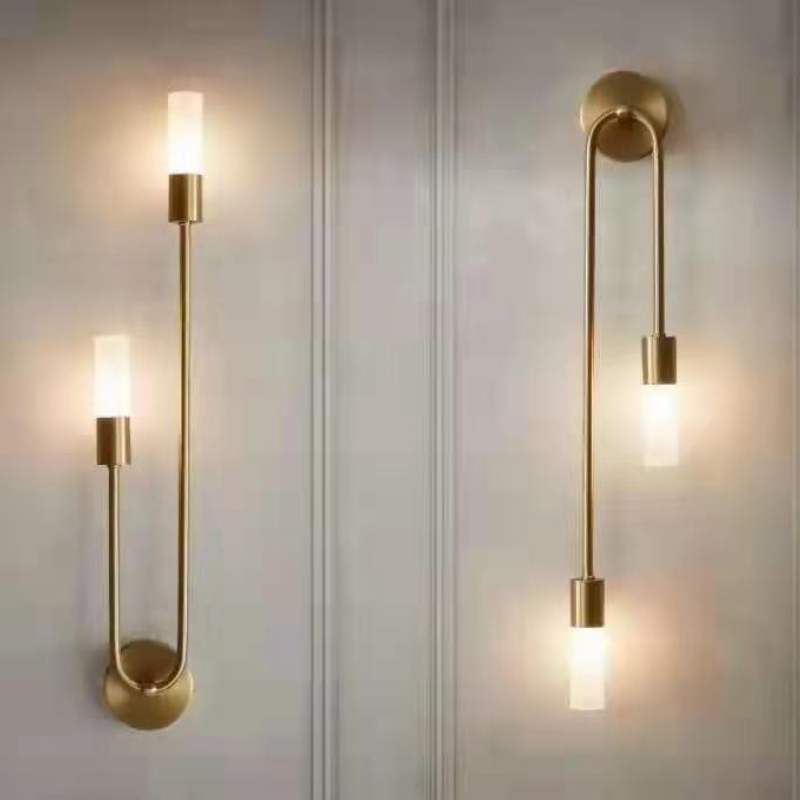 Two Clérachic gold stick sconces staggered on a panelled wall, both frosted shades lit warm white.