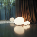 Clérachic glowing opal glass egg lights in three sizes on a dark glossy floor in front of a copper-toned curtain.