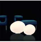 Clérachic two lit opal glass egg lights in different sizes on a dark floor in front of blue upholstered armchairs.