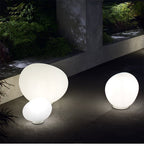 Clérachic three lit opal glass egg lights of different sizes on a paved terrace beside a concrete wall and green planting.