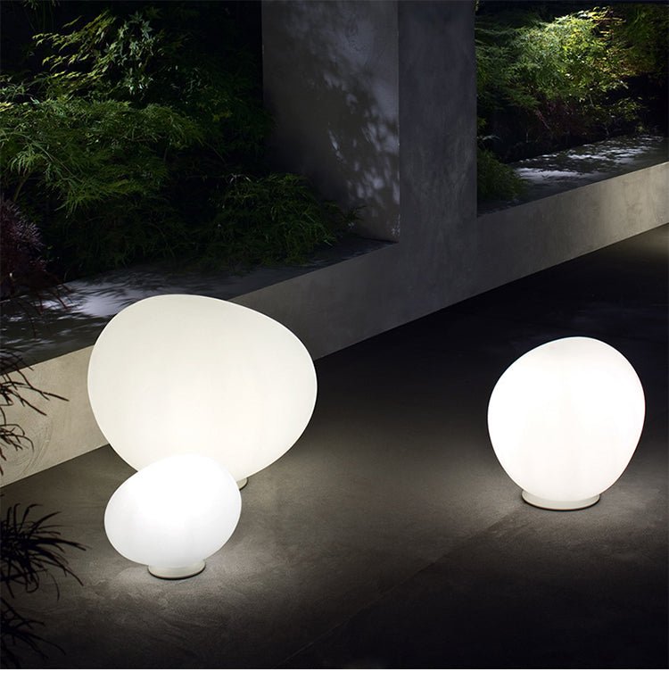 Clérachic three lit opal glass egg lights of different sizes on a paved terrace beside a concrete wall and green planting.