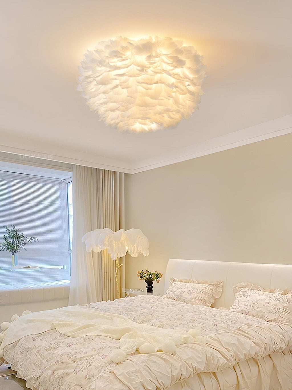 White feather ceiling light from Clérachic lit above a cream bedroom with a ruffled bed, a feather floor lamp and soft curtains.
