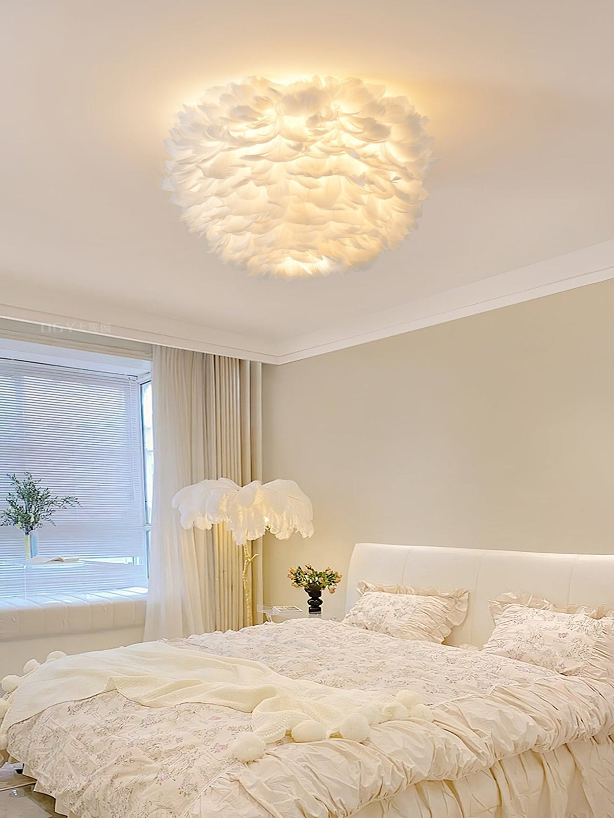 White feather ceiling light from Clérachic lit above a cream bedroom with a ruffled bed, a feather floor lamp and soft curtains.