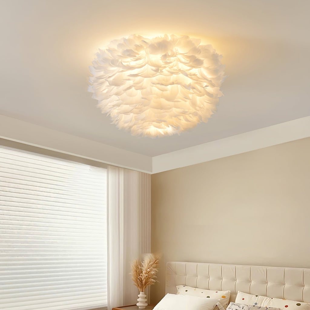 Clérachic feather ceiling light glowing above a bedroom corner with a cream headboard, a roller blind and a vase of pampas grass.