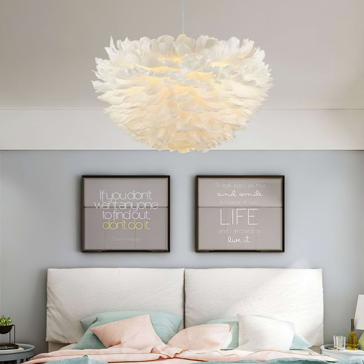 A Clérachic pendant with a fluffy white feather dome hangs above a bed with pastel cushions and two framed text prints.