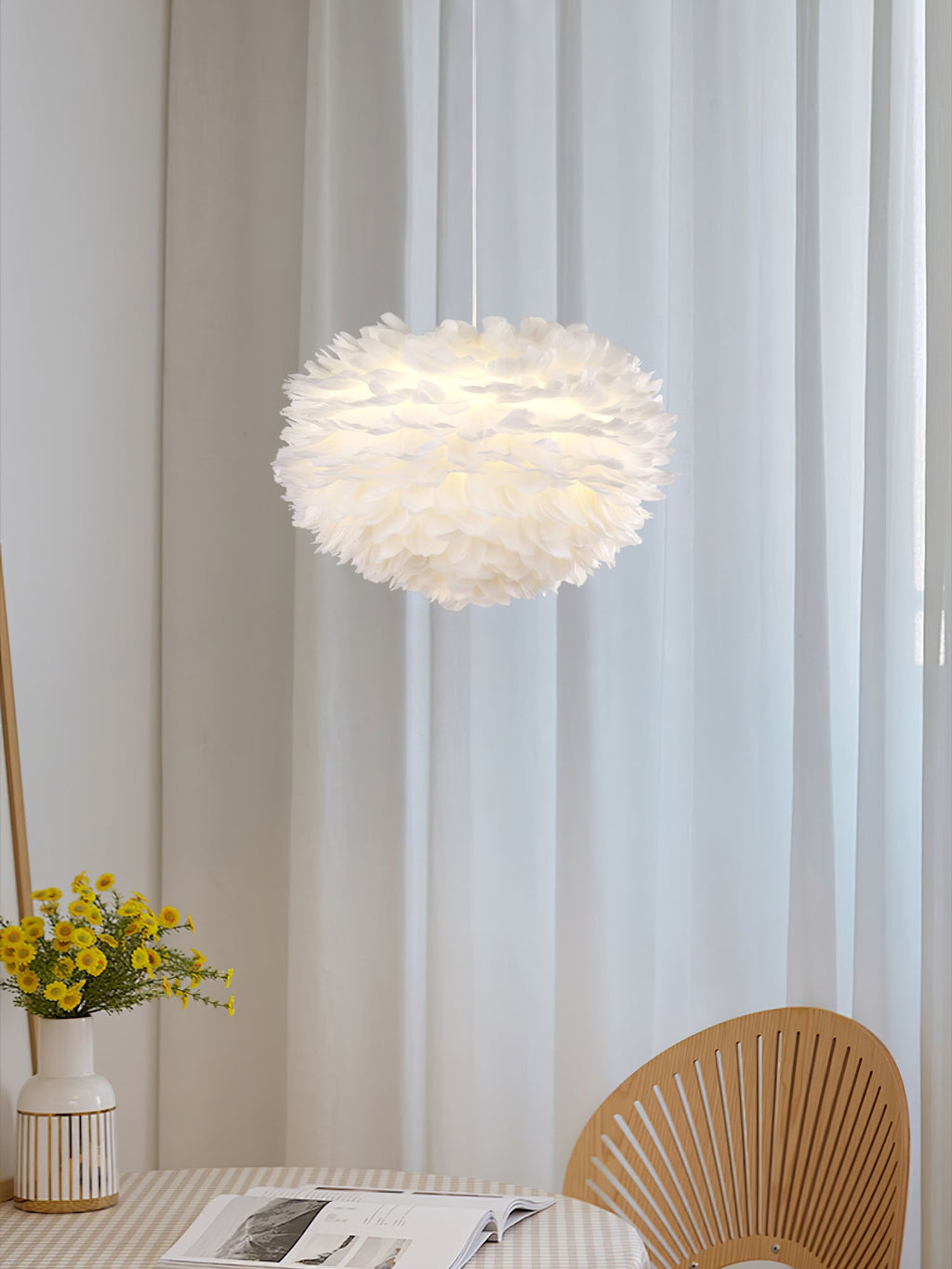 A small Clérachic feather pendant hangs between sheer curtains, with yellow flowers and a rattan fan-back chair below.