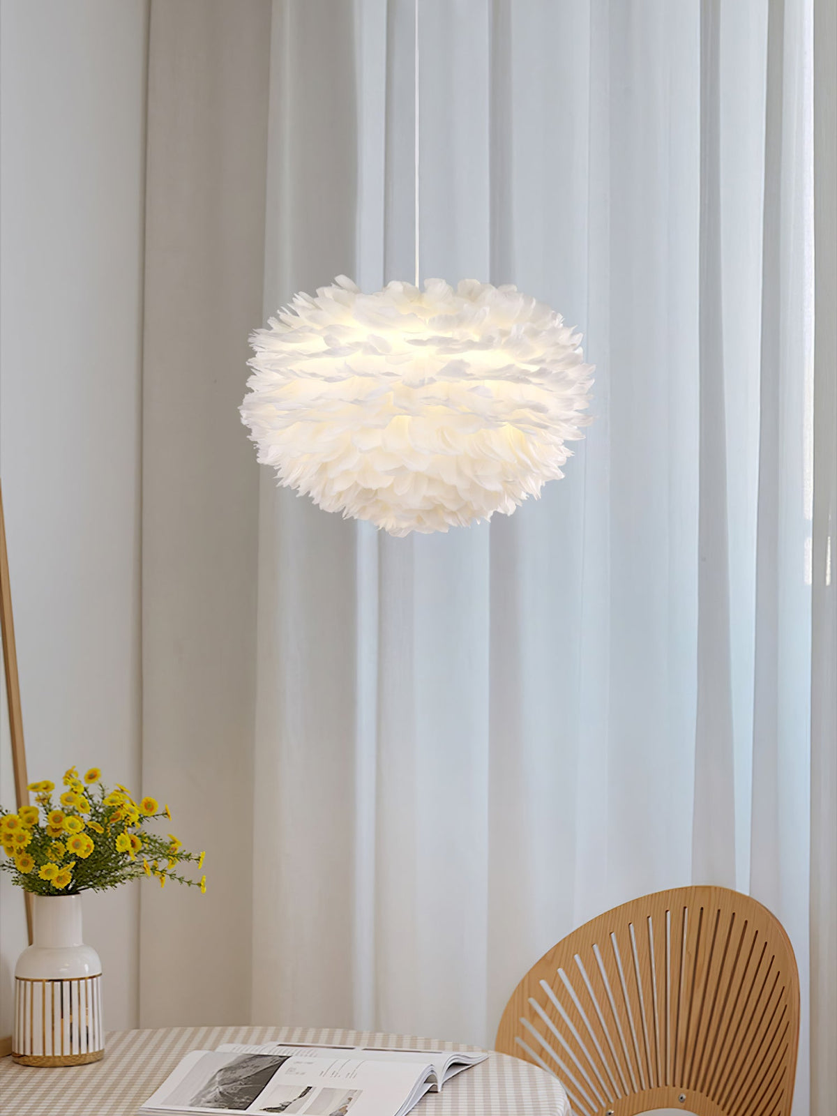 A small Clérachic feather pendant hangs between sheer curtains, with yellow flowers and a rattan fan-back chair below.