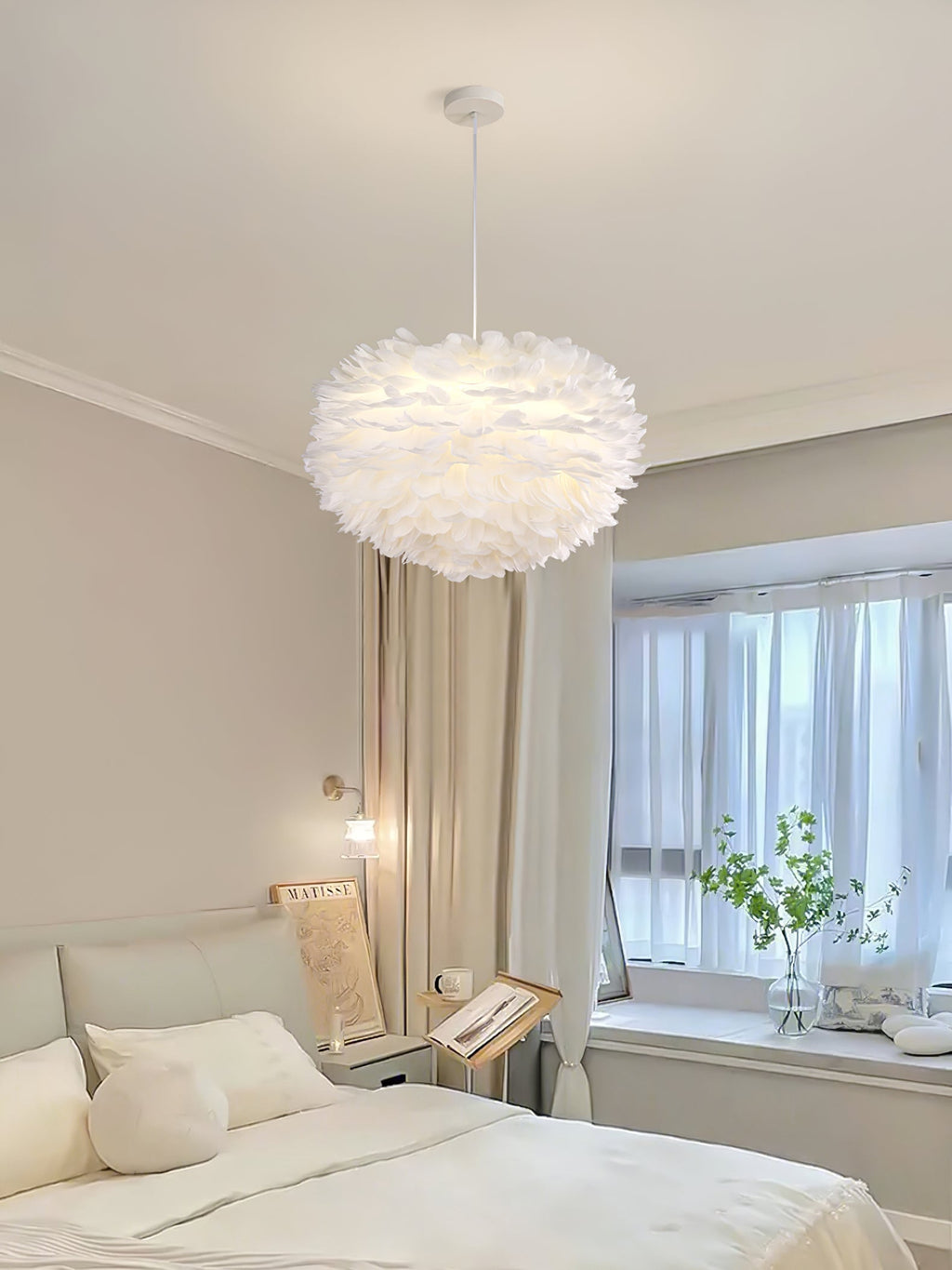 A white dome Clérachic feather pendant hangs in a calm bedroom, with a window, a pale bed and a green plant nearby.