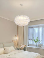 A white dome Clérachic feather pendant hangs in a calm bedroom, with a window, a pale bed and a green plant nearby.