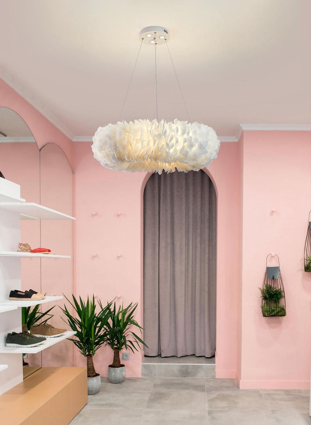 A Clérachic pendant with a wide ring of feathers hangs in a pink-walled shop, with shelving and plants beside an arched curtain.