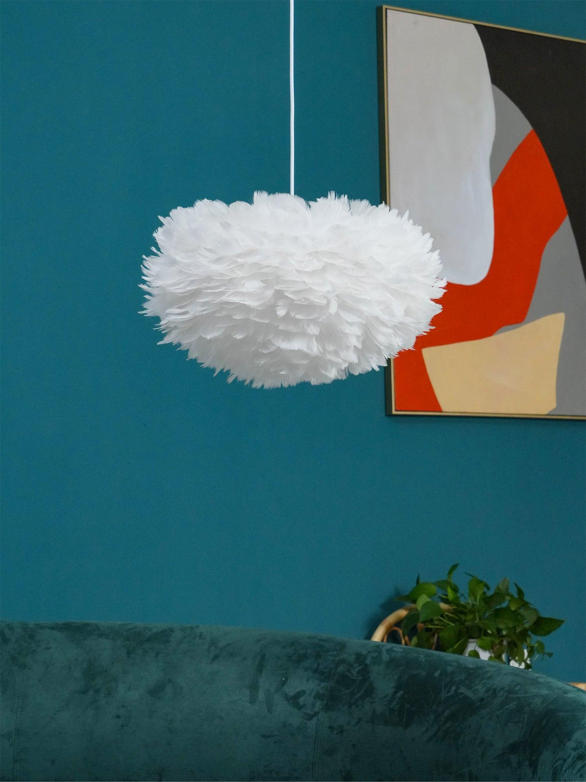 A Clérachic feather pendant hangs against a deep teal wall beside an abstract artwork, with a green velvet sofa below.