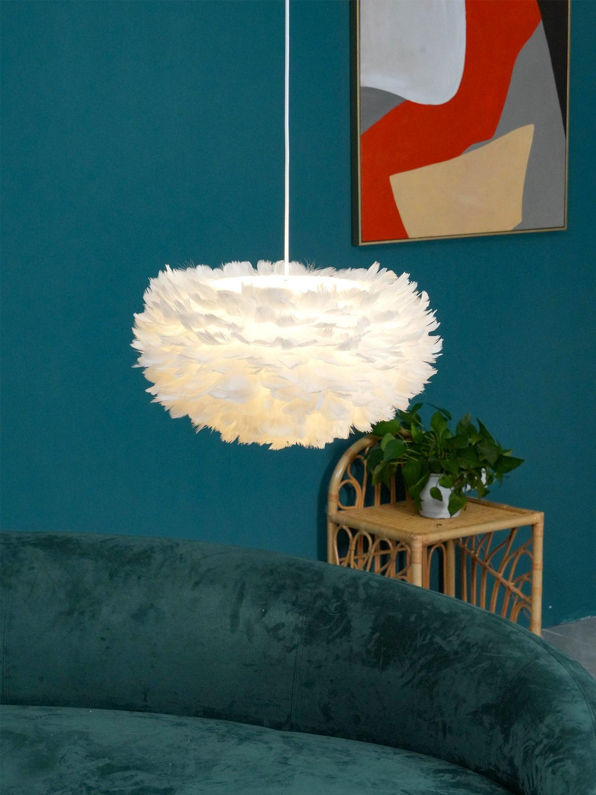 Another view of the Clérachic feather pendant in front of a teal wall, with a rattan side table and a leafy plant below.