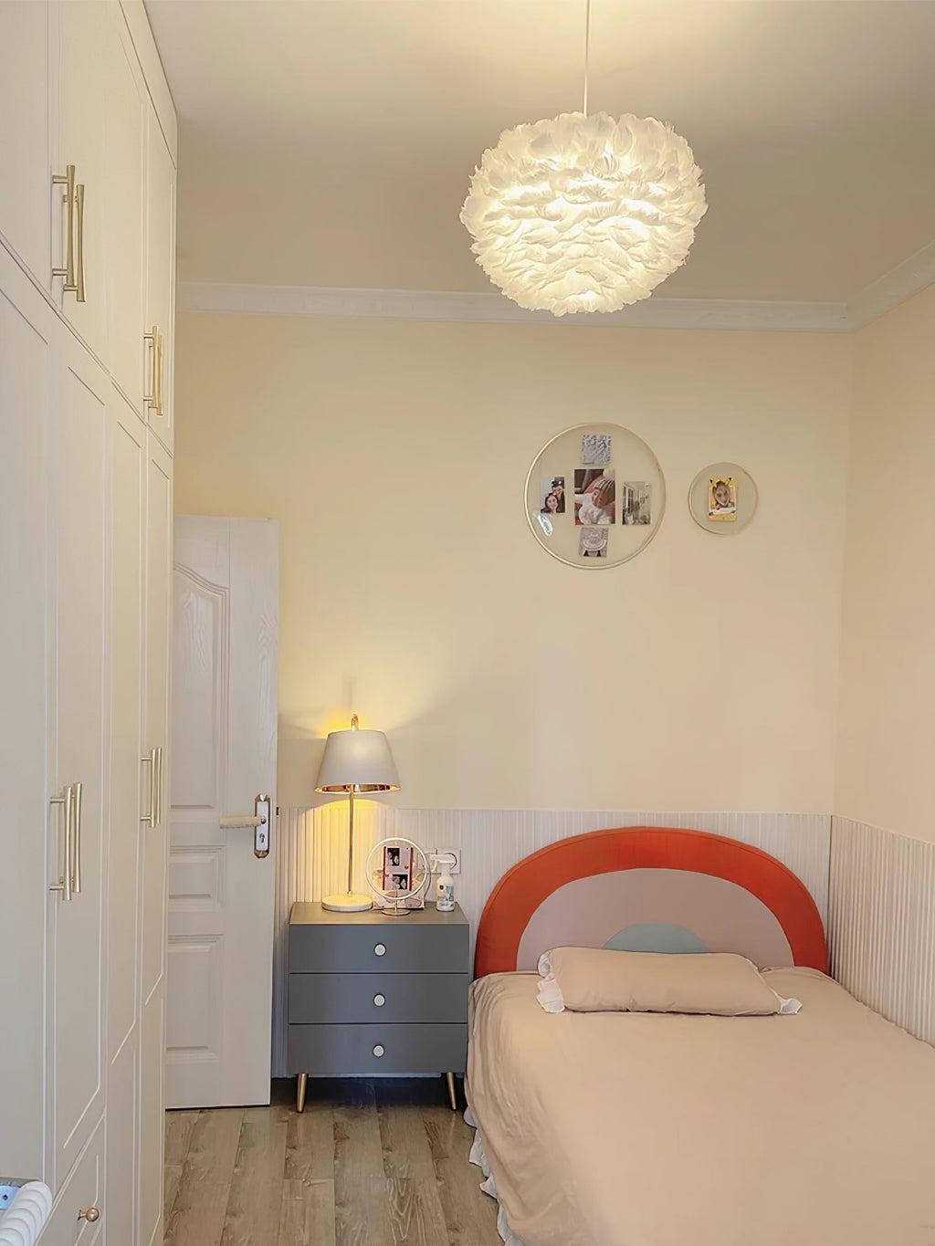 A Clérachic feather pendant glows in a cream child's bedroom with a coral headboard, a grey bedside chest and a lamp.