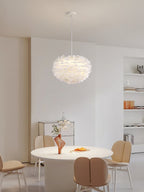 A Clérachic feather pendant hangs above a round white dining table with beige chairs, in a bright room with open shelves.