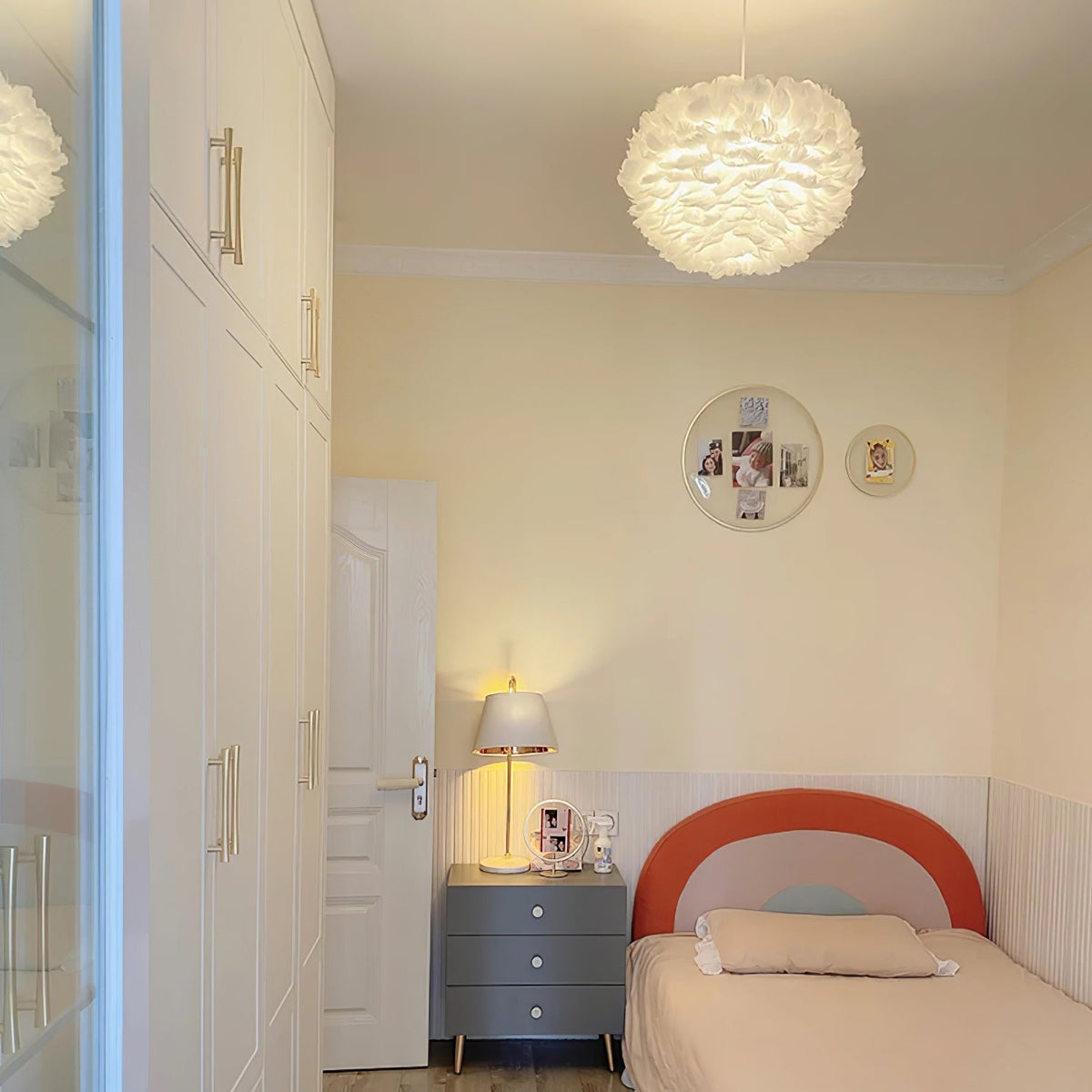 A Clérachic feather pendant lit above a child's bed with a coral headboard, in a room with cream walls and a bedside lamp.